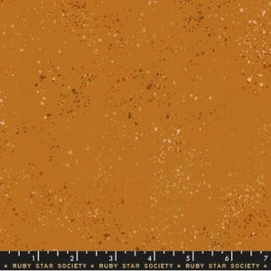 May include: A mustard yellow fabric with a speckled pattern of brown, gold, and pink dots. The fabric is a solid color with a subtle texture.