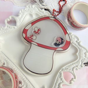 May include: A clear mushroom-shaped keychain with a red and white polka dot border. Inside are small mushroom and character illustrations. A red beaded chain is attached. Decorative tape rolls with watermelon designs are also visible.