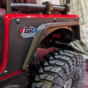 Puede incluir: Un conjunto de guardabarros rojo para veh&iacute;culo todoterreno con un protector negro texturizado, una pegatina de GenRight Off Road y torniller&iacute;a visible. El componente est&aacute; fijado a un panel rojo sobre un neum&aacute;tico negro de gran relieve.