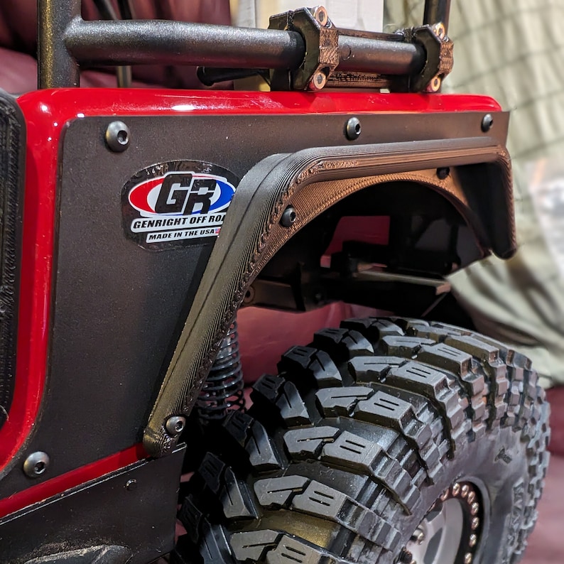 Puede incluir: Un conjunto de guardabarros rojo para veh&iacute;culo todoterreno con un protector negro texturizado, una pegatina de GenRight Off Road y torniller&iacute;a visible. El componente est&aacute; fijado a un panel rojo sobre un neum&aacute;tico negro de gran relieve.
