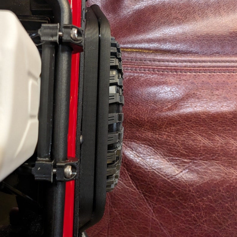 Puede incluir: Un protector de guardabarros de pl&aacute;stico negro montado en un chasis de veh&iacute;culo rojo, con un acabado texturizado y fijado con tornillos met&aacute;licos visibles. El componente est&aacute; colocado junto a un neum&aacute;tico de goma negro con banda de rodadura.