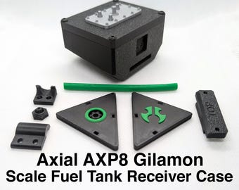 Puede incluir: Una caja receptora de tanque de combustible a escala Axial AXP8 Gilamon con una caja negra texturizada, una placa superior plateada y varios componentes de plástico negro y verde, incluyendo piezas triangulares, un tubo verde y una pieza rectangular etiquetada como TOP.