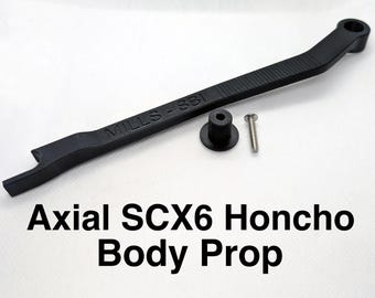 Pode incluir: Um suporte de carroçaria preto Axial SCX6 Honcho com o texto MILLS - SBI em relevo na superfície. O conjunto inclui um componente preto longo e fino com uma extremidade circular, um pequeno espaçador cilíndrico preto e um parafuso metálico prateado.
