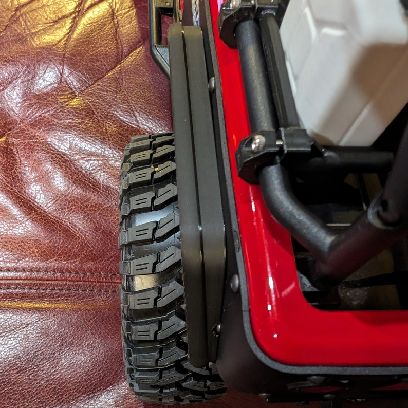 Puede incluir: Una vista de cerca de un veh&iacute;culo a control remoto rojo que presenta un neum&aacute;tico negro texturizado con bandas de rodadura profundas, un panel lateral negro con remaches met&aacute;licos visibles y una estructura de jaula antivuelco tubular negra junto a un componente de pl&aacute;stico blanco.