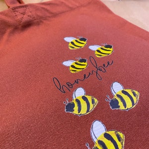 May include: A red canvas tote bag with the word "honeybee" written in cursive and six yellow and black bee illustrations.