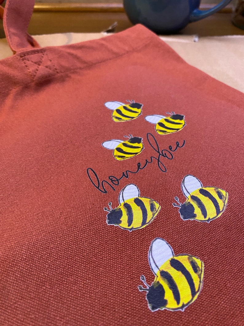 May include: A red canvas tote bag with the word "honeybee" written in cursive and six yellow and black bee illustrations.