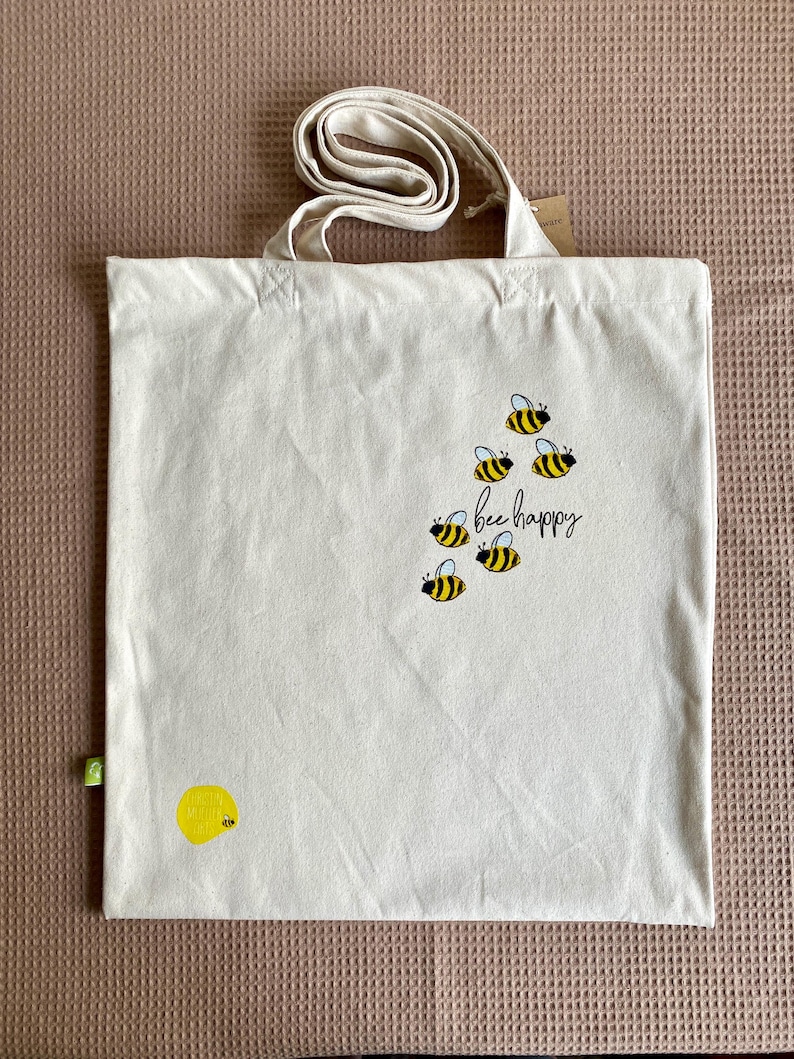 May include: A white canvas tote bag with a yellow bee design and the text "bee happy".