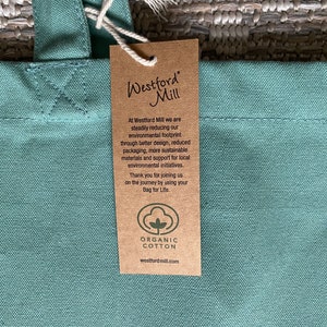 May include: A green canvas tote bag with a brown paper tag attached. The tag has the text "Westford Mill" and information about the company's commitment to sustainability and using organic cotton. The tag also includes the company's website address: westfordmill.com.