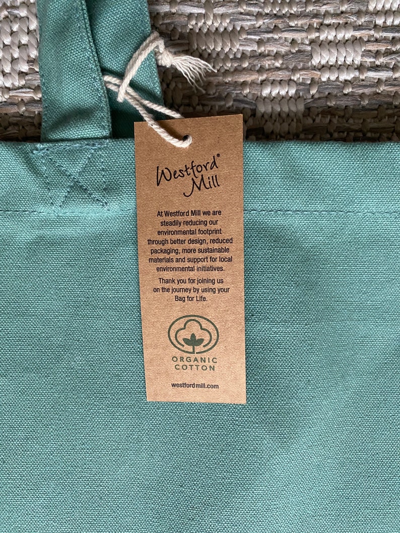 May include: A green canvas tote bag with a brown paper tag attached. The tag has the text "Westford Mill" and information about the company's commitment to sustainability and using organic cotton. The tag also includes the company's website address: westfordmill.com.