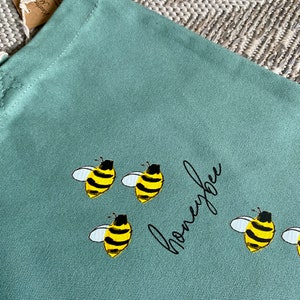 May include: A light green canvas tote bag with the word "honeybee" written in black cursive script. There are five yellow and black bees with white wings printed on the bag.