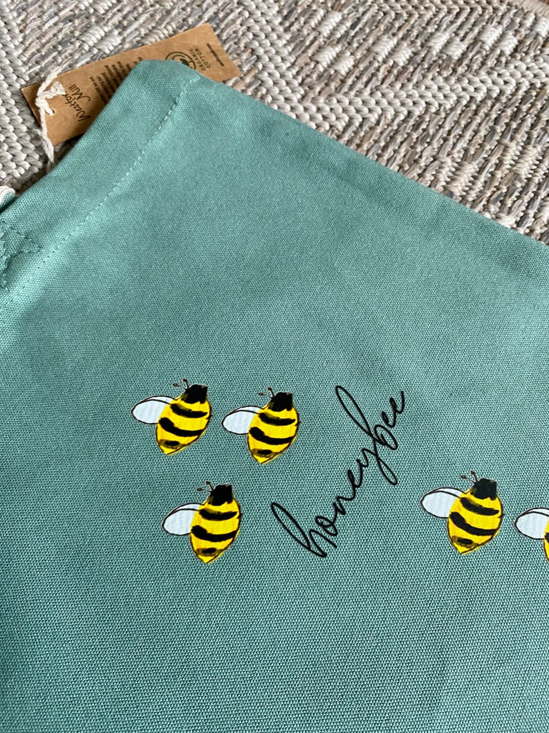 May include: A light green canvas tote bag with the word "honeybee" written in black cursive script. There are five yellow and black bees with white wings printed on the bag.
