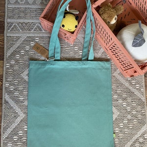 May include: A light teal canvas tote bag with two handles. The bag is sitting on a patterned rug with two pink plastic storage bins in the background.