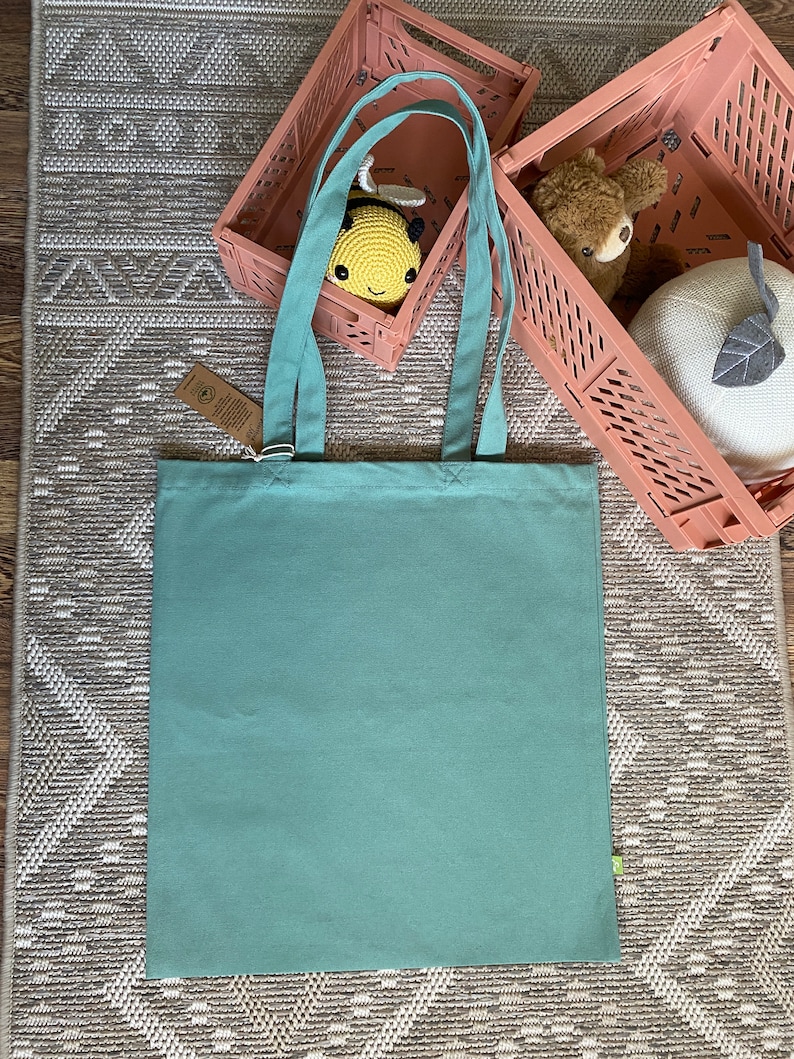 May include: A light teal canvas tote bag with two handles. The bag is sitting on a patterned rug with two pink plastic storage bins in the background.
