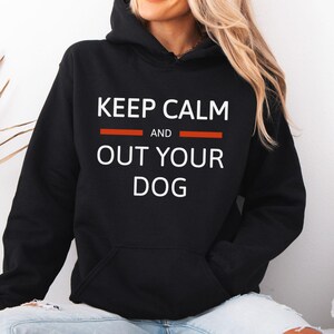 May include: A black hooded sweatshirt featuring white text that reads KEEP CALM AND OUT YOUR DOG, with two horizontal orange lines flanking the word AND. The garment includes a front kangaroo pocket and long sleeves with ribbed cuffs.