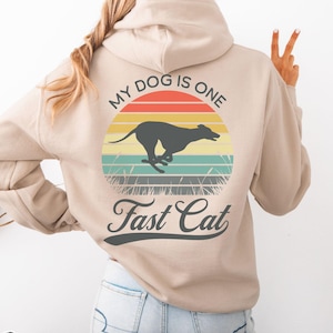 May include: Sand-colored hoodie with a graphic on the back featuring a running dog silhouette against a sunset design. The text reads "My Dog Is One Fast Cat". The hoodie is made of a soft material.