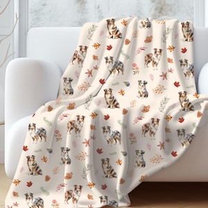 May include: A cream-colored throw blanket featuring a repeating pattern of Australian Shepherd dogs and autumn leaves in shades of brown, red, and green. The blanket is draped over a white sofa.