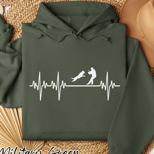 May include: An olive green hooded sweatshirt featuring a white graphic print of a heartbeat line, a jumping dog, and a person standing. The hoodie includes a drawstring hood, long sleeves, and a front kangaroo pocket in a solid dark green color.
