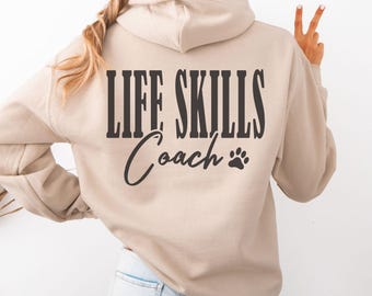 Puede incluir: Una sudadera con capucha color arena con el texto "LIFE SKILLS Coach" impreso en negro en la espalda. Un gráfico de huella de pata está debajo de la palabra "Coach". La sudadera tiene capucha y mangas largas.