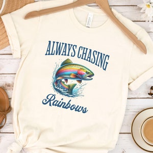 May include: A white t-shirt with the text "ALWAYS CHASING RAINBOWS" in blue lettering. A rainbow trout jumps out of the water, surrounded by blue waves.