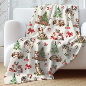 May include: A cream-colored throw blanket featuring a repeating pattern of Australian Shepherds, Christmas trees, wrapped gifts, and red bows. Some dogs wear Santa hats. The blanket is draped over a white sofa.