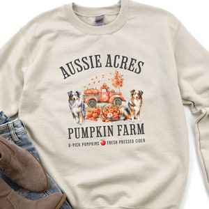 May include: A beige sweatshirt with a graphic design of two Australian Shepherds standing in front of a red truck with pumpkins. The text "Aussie Acres Pumpkin Farm" is above the truck and "U-Pick Pumpkins Fresh Pressed Cider" is below the truck.