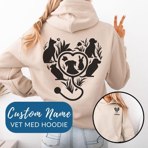 Peut inclure: Un sweat à capuche beige avec un graphique noir d'un stéthoscope et d'animaux en forme de cœur au dos. Le sweat à capuche a une poche sur le devant et le texte "Custom Name VET MED HOODIE" est imprimé sous le graphique. Une image plus petite du sweat à capuche montre le texte "Ashley" imprimé au dos.