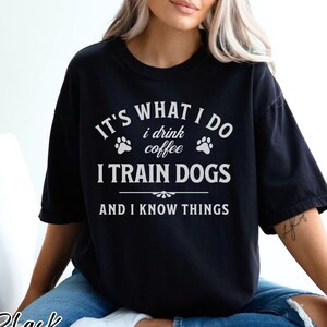May include: Black t-shirt with white text: "IT'S WHAT I DO, I DRINK COFFEE, I TRAIN DOGS, AND I KNOW THINGS." The shirt has a relaxed fit and is made of a soft material. The word "Black" is printed at the bottom left.
