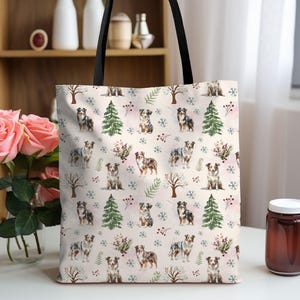 May include: A cream-colored tote bag with a black handle features a repeating pattern of Australian Shepherd dogs, snowflakes, trees, and berries. The bag is made of a sturdy fabric and is suitable for carrying groceries or personal items.