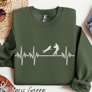 May include: Military green sweatshirt with a white graphic of a dog and person, with a heartbeat-like line. The text "Military Green" is at the bottom. The sweatshirt has a crew neck and long sleeves.