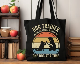 May include: Black tote bag with a retro-style design featuring a dog and a person. The text reads "Dog Trainer Making the World a Better Place One Dog at a Time."