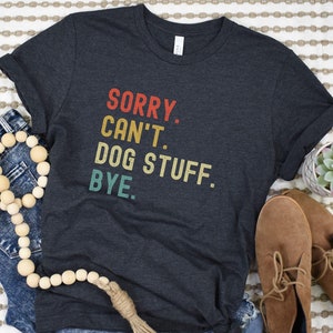 以下が含まれることがあります： 「SORRY. CAN'T. DOG STUFF. BYE.」とヴィンテージスタイルのグラフィックがプリントされたダークグレーのTシャツです