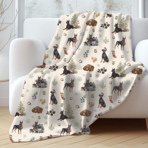 May include: A cream-colored throw blanket featuring a repeating pattern of Doberman Pinscher dogs, Christmas trees, wrapped gifts, pinecones, snowflakes, and stockings. Some dogs are wearing Santa hats, while others are resting near holiday decor.