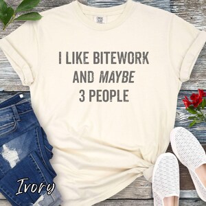 May include: Ivory-colored t-shirt with the text "I LIKE BITWORK AND MAYBE 3 PEOPLE" in gray. The shirt is paired with blue jeans and white striped shoes. A sprig of red flowers and green leaves are also visible.
