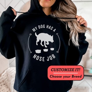May include: Black hoodie with a white graphic that says "My dog has a nose job" with a silhouette of a dog doing a nose work exercise. The text "Customize it! Choose your breed" is in a red box at the bottom of the hoodie.