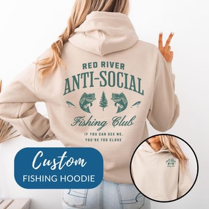 May include: A beige hoodie with a green graphic on the back that says "Red River Anti-Social Fishing Club" and "If you can see me, you're too close". The hoodie has a hood and a kangaroo pocket. The hoodie is shown on a model wearing blue jeans.