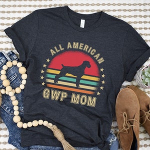 May include: A dark grey t-shirt with a vintage-style graphic of a German Shorthaired Pointer dog in front of a sunset. The text "All American GWP Mom" is printed below the graphic.