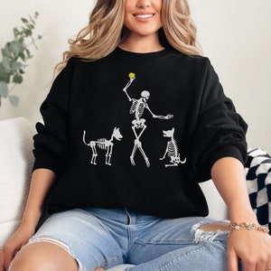 May include: A black sweatshirt with a white skeleton graphic of a person playing fetch with two dogs. The skeleton is holding a yellow ball in its hand.