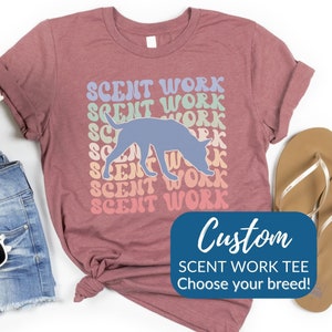 May include: A maroon t-shirt with the text "SCENT WORK" repeated in a retro style, with a light blue dog silhouette. The shirt also has a blue rectangle with the text "Custom Scent Work Tee Choose your breed!"