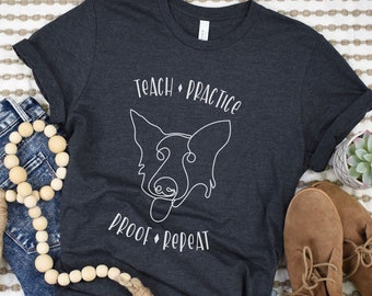 May include: A dark gray t-shirt with a white outline of a dog's head and the text "Teach Practice Proof Repeat".