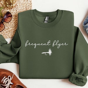 May include: A dark green sweatshirt with the text "frequent flyer" in white script and a white silhouette of a fishing fly.