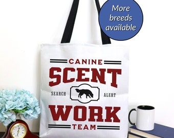 May include: White canvas tote bag with black handles. The bag has a red and black graphic that says "Canine Scent Work Team" with a silhouette of a dog in the middle. The text "Search" and "Alert" are on either side of the dog silhouette.
