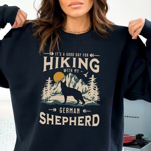 May include: Black sweatshirt with a graphic design featuring a silhouette of a German Shepherd dog in a mountain landscape. The text reads "It's a good day for hiking with my German Shepherd".