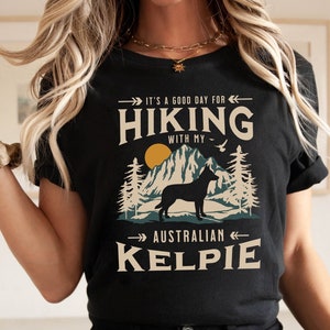 May include: Black t-shirt with a graphic design featuring a silhouette of an Australian Kelpie dog hiking in a mountain landscape. The text reads "It's a good day for hiking with my Australian Kelpie."