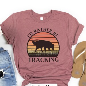 May include: Heather mauve t-shirt with a retro sunset graphic and a silhouette of a dog. The text "I'd rather be tracking" is printed below the graphic.