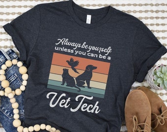 May include: Dark grey heather t-shirt with a retro-style graphic that says "Always be yourself unless you can be a Vet Tech". The graphic features a cat, dog, and bird in front of a sunset.