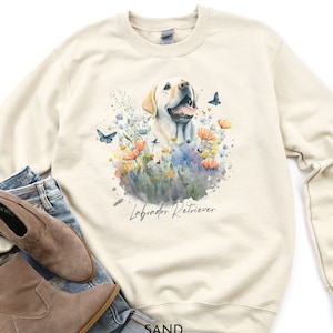 May include: A cream-colored sweatshirt with a watercolor illustration of a yellow Labrador Retriever surrounded by flowers and butterflies. The text "Labrador Retriever" is printed below the dog.