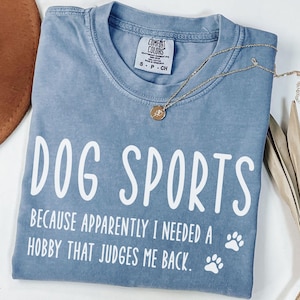 May include: A blue Comfort Colors t-shirt with the text "DOG SPORTS" and the phrase "BECAUSE APPARENTLY I NEEDED A HOBBY THAT JUDGES ME BACK." Two white paw prints are below the text. A gold necklace is draped across the shirt.