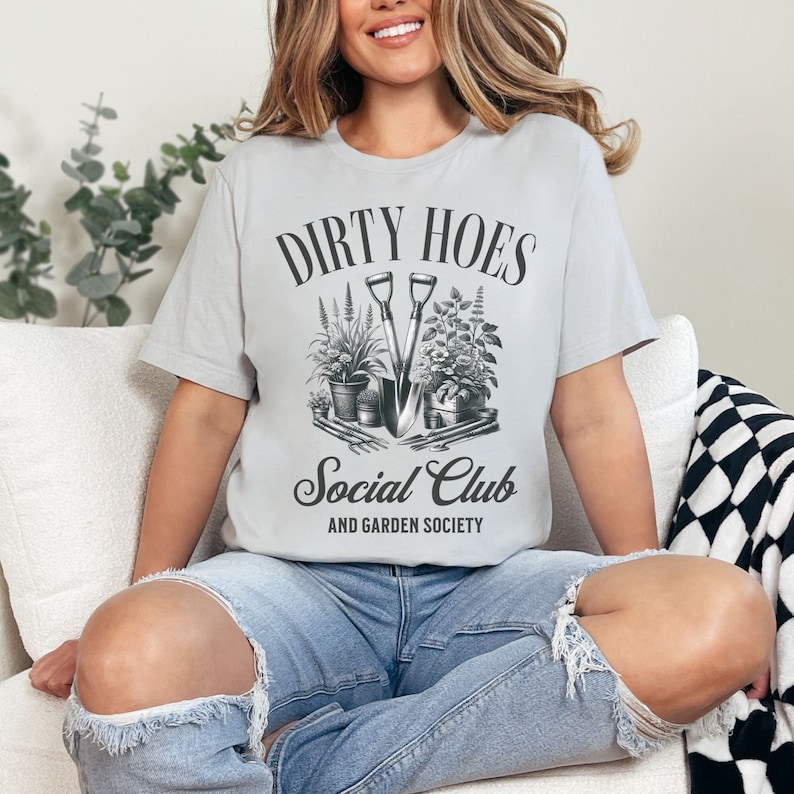 May include: A gray t-shirt with the text "Dirty Hoes Social Club and Garden Society" in black lettering. The design features a pair of gardening tools and plants.