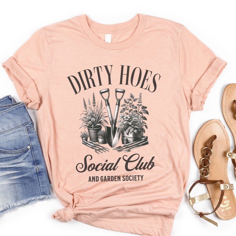 May include: A peach-colored t-shirt with the text "Dirty Hoes Social Club and Garden Society" in black. The design features a pair of gardening tools and plants.
