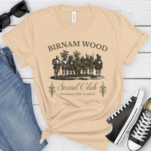 May include: A beige t-shirt with a graphic of a group of men dressed as trees, with the text "Birnam Wood Social Club Dunsinane Hill, Scotland".